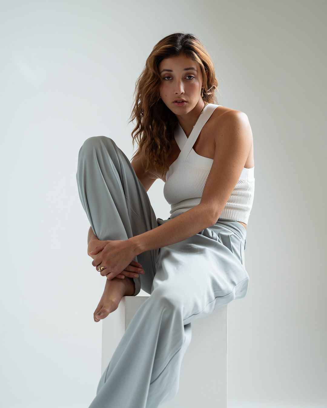 Person in white halter top and light gray pants poses against white wall in contemporary minimalist portrait.