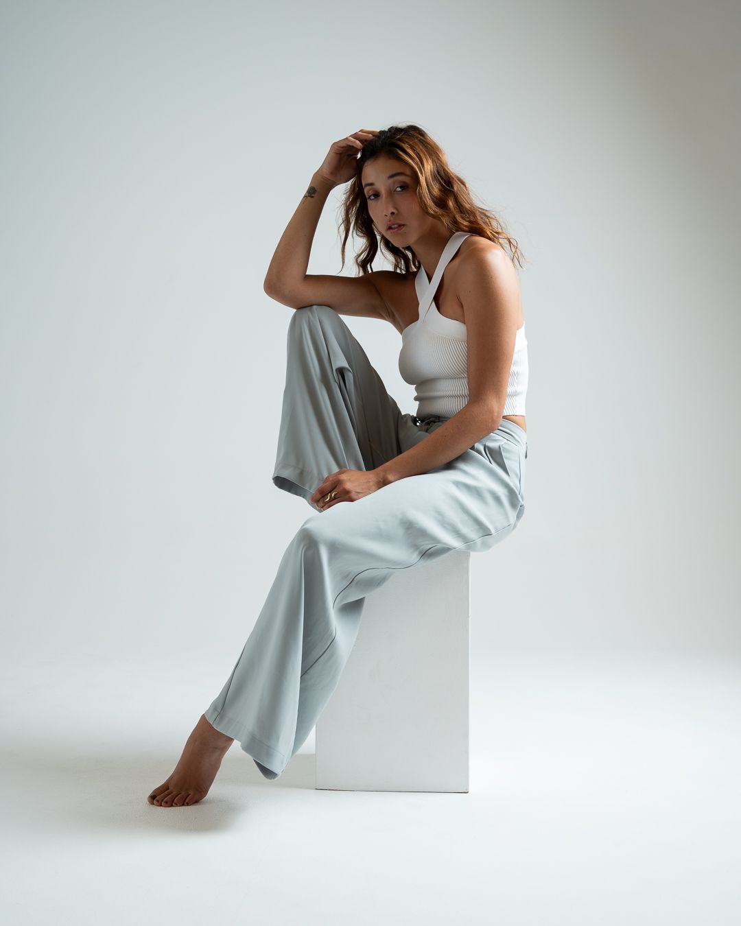 Artistic portrait of model in casual white and gray outfit seated on minimalist white pedestal.