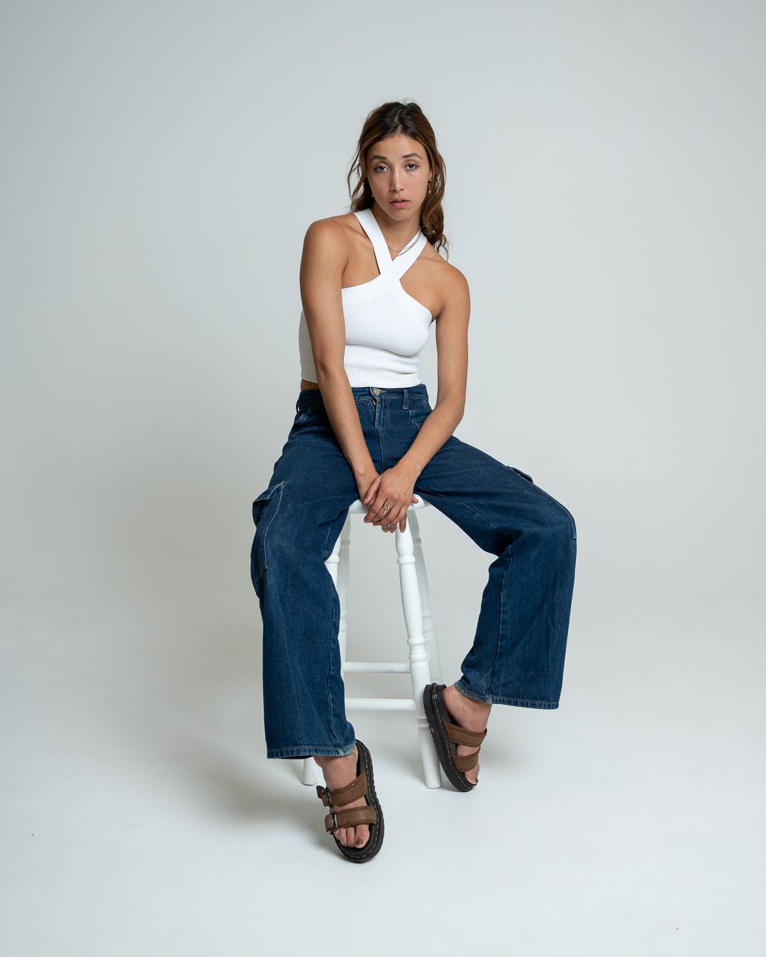 A person in a white tank top and wide-leg jeans sits casually on a white chair against a light gray background.