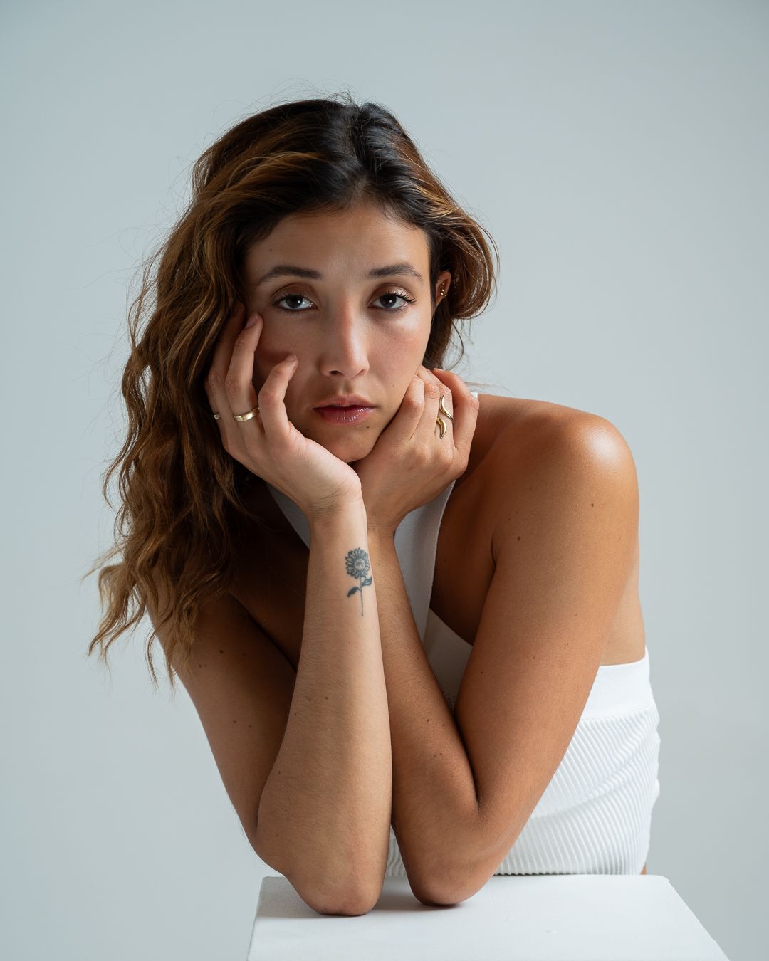A woman in a white top poses with hands resting on face against a light gray background.