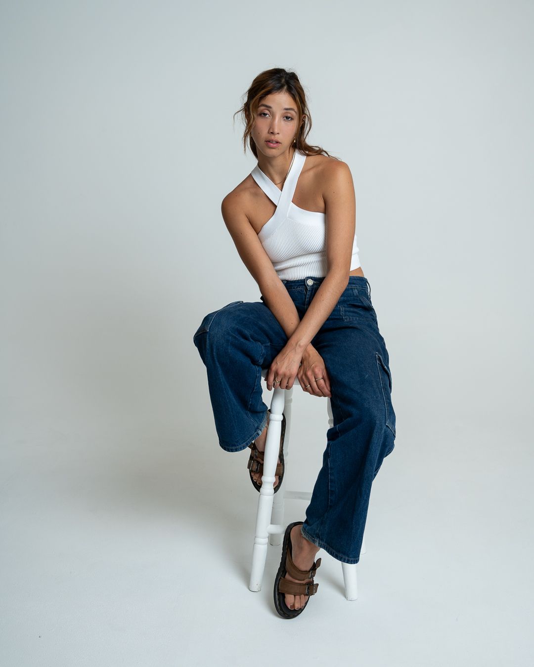 A person in casual attire poses on a white chair in a minimalist studio setting with soft lighting.