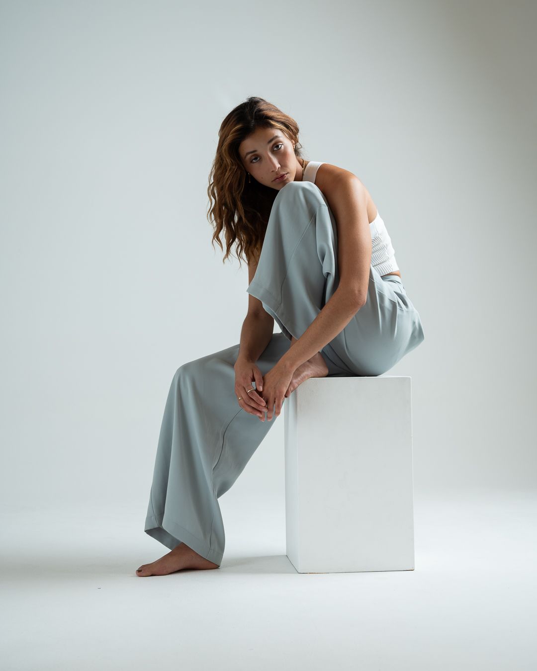 Fashion portrait of someone in a light blue outfit sitting elegantly on a white pedestal against neutral background.