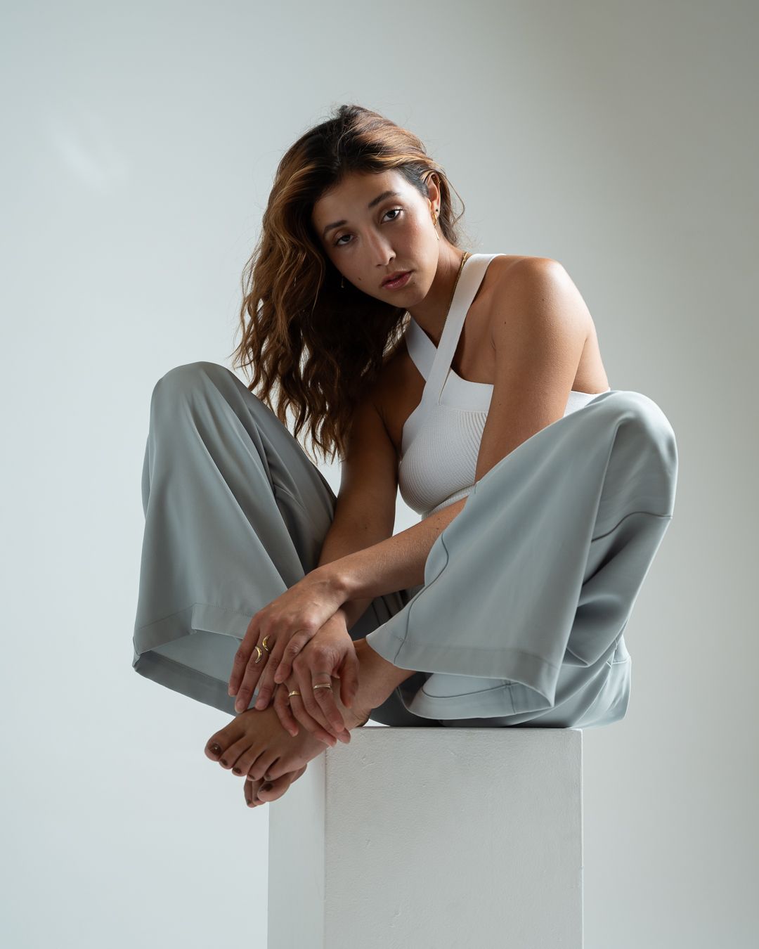 Fashion model in white tank top and light gray pants poses elegantly on white cube against neutral background.