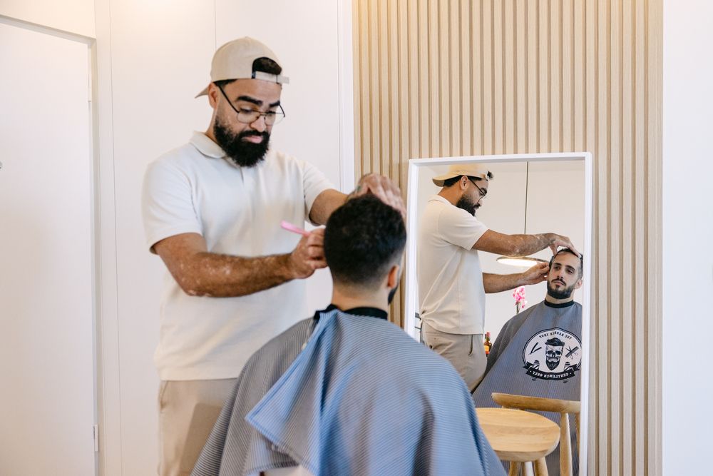 Professional barber in white t-shirt and cap cutting groom's hair in modern house with mirror reflection.