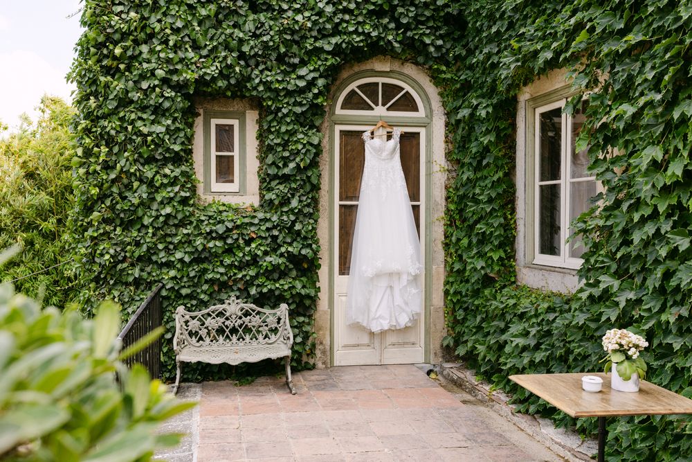 A white wedding dress hangs in an elegant doorway of an ivy-covered stone building with a decorative bench nearby in solar de pancas