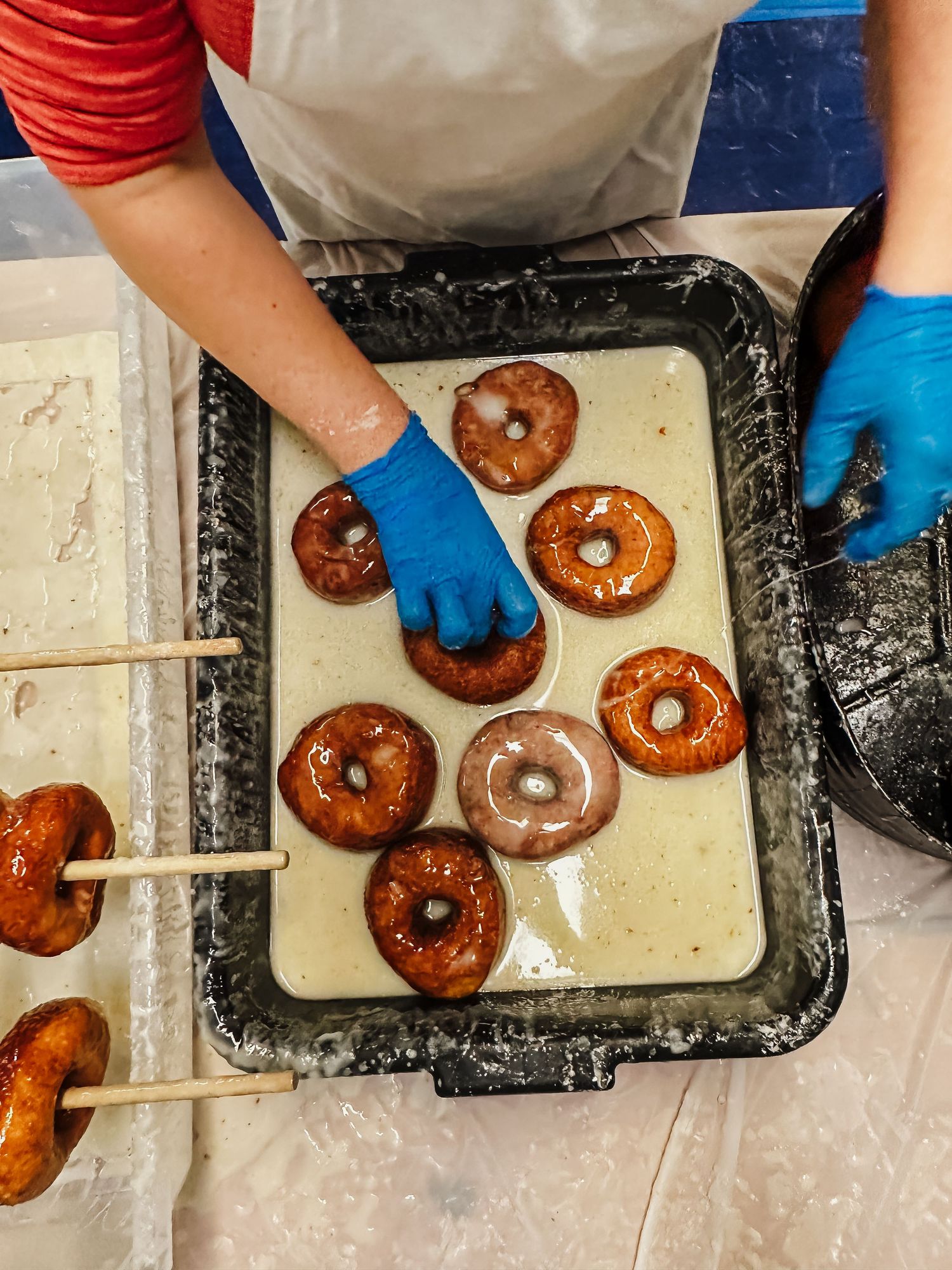 Hands wearing blue gloves dip fresh glazed donuts in a tray filled with coating mixture.