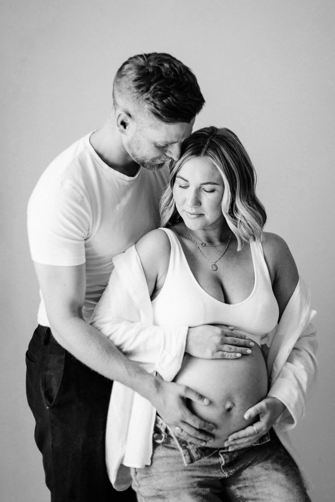 Black and white maternity photo showing intimate moment between expecting couple in white clothing and denim.