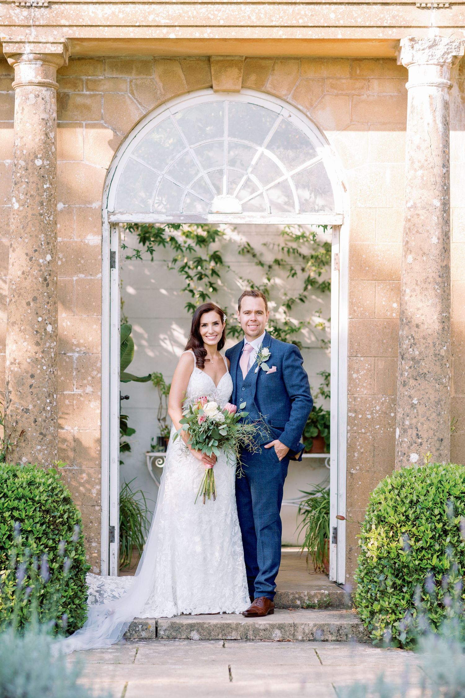 Newlywed couple poses together in elegant stone archway with climbing vines at romantic garden wedding venue.