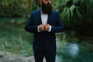 A person in a navy suit stands in front of a lush green garden setting with a flowing stream in the background.
