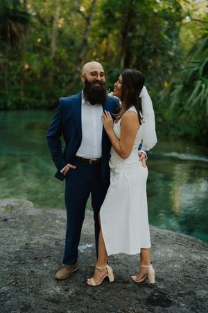 A couple shares an intimate moment by a turquoise lagoon, with the bride in white and groom in navy blue suit.