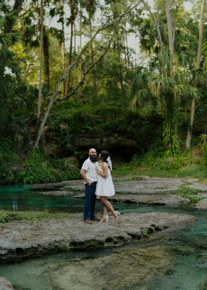 A couple sits together on rocky terrain by a natural spring pool surrounded by lush tropical palm trees and foliage.