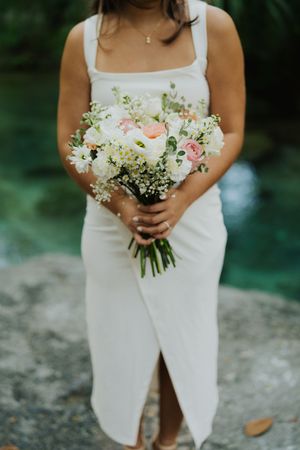A close-up detail shot of a simple white wedding dress and bridal bouquet with pink and white flowers against a green background.