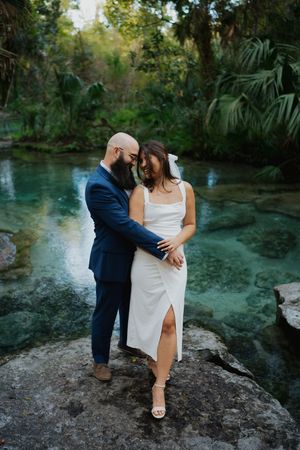 A romantic couple embraces on a rock overlooking crystal clear turquoise waters surrounded by lush tropical foliage.