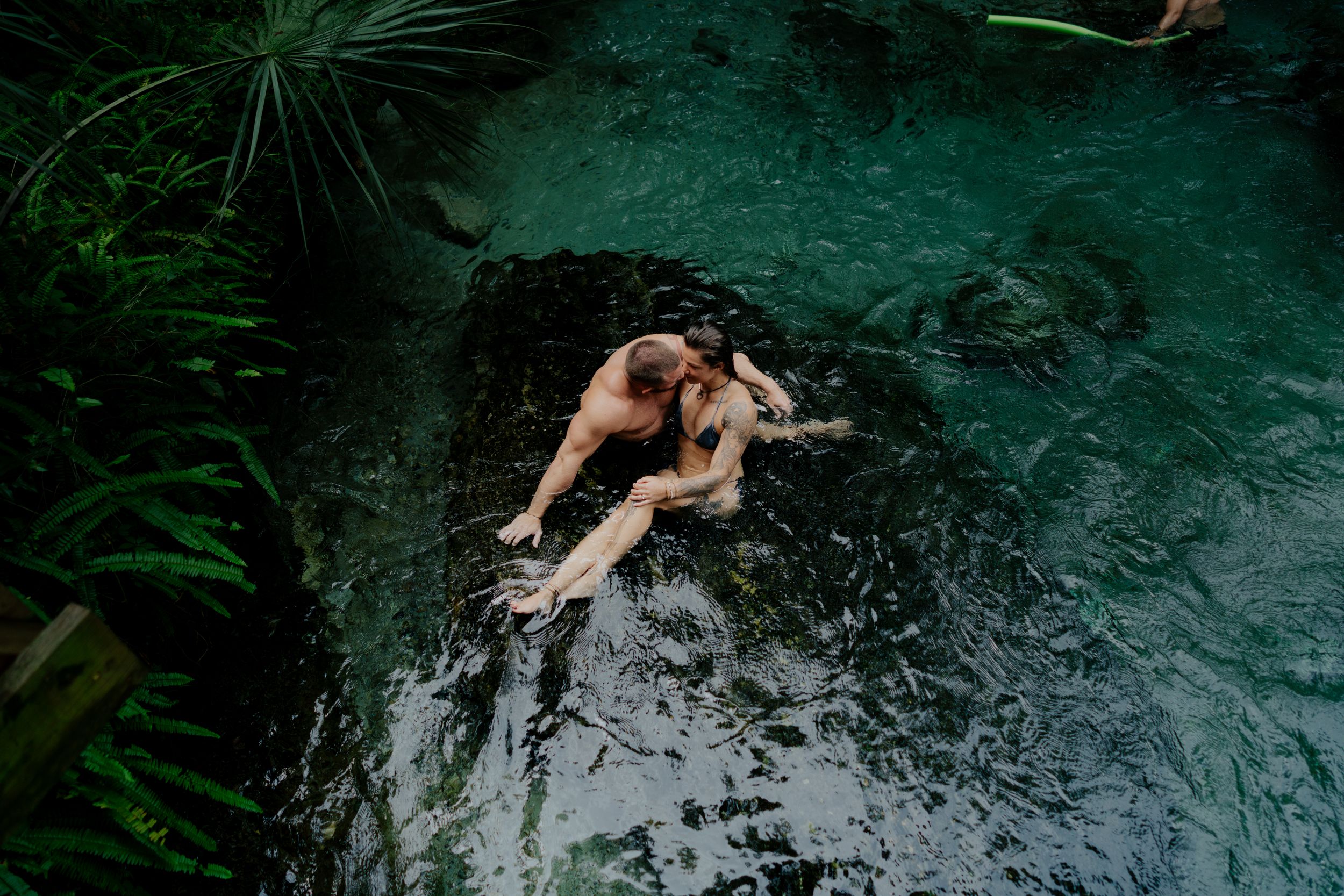 Couple swimming in crystal clear turquoise cenote waters surrounded by lush vegetation and natural rock formations.