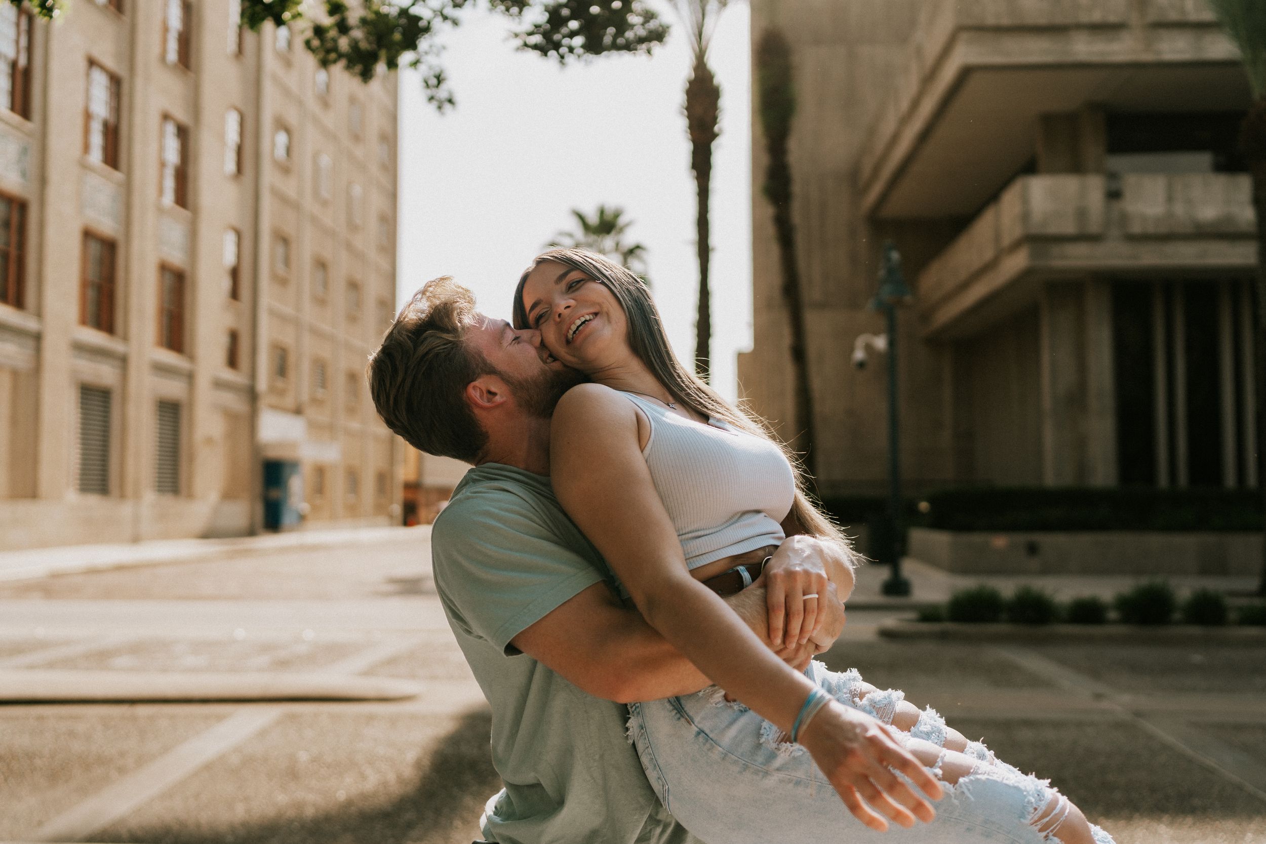A couple embraces in a romantic moment on an urban street corner with warm sunlit architecture in the background.