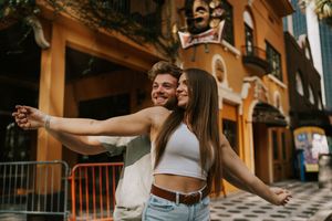 A joyful couple spreads their arms wide while standing in front of a yellow Mediterranean-style building.