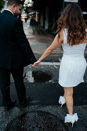 Couple in wedding attire walking together on wet pavement outside a green building.