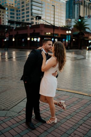 Couple embracing on brick pavement with city buildings in the background at dusk.
