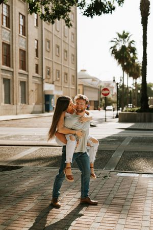 A playful couple shares joyful moments together on a paved walkway with buildings in the background.