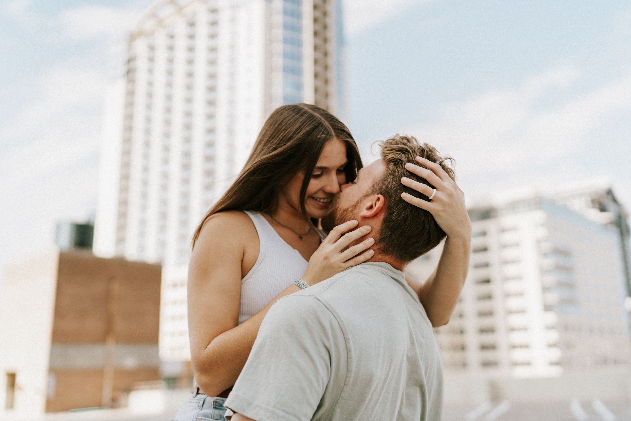 A romantic couple shares an intimate moment on a rooftop with a cityscape backdrop and tall buildings behind them.