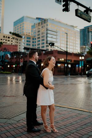 A couple shares a joyful moment together on a rainy city street with urban buildings in the background.