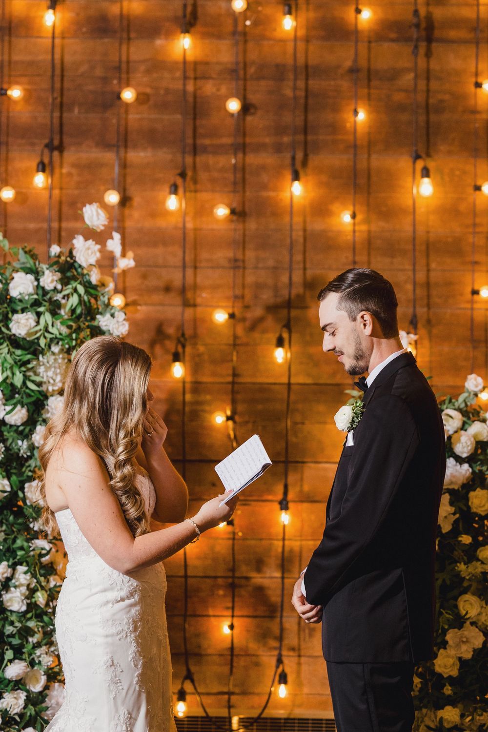 A couple shares a private moment reading letters to each other in front of a rustic wood wall with glowing string lights.