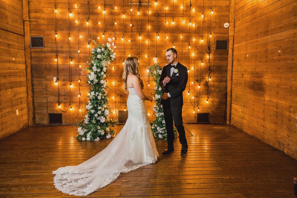 A couple walks through a rustic wooden barn venue decorated with warm string lights and Christmas trees.