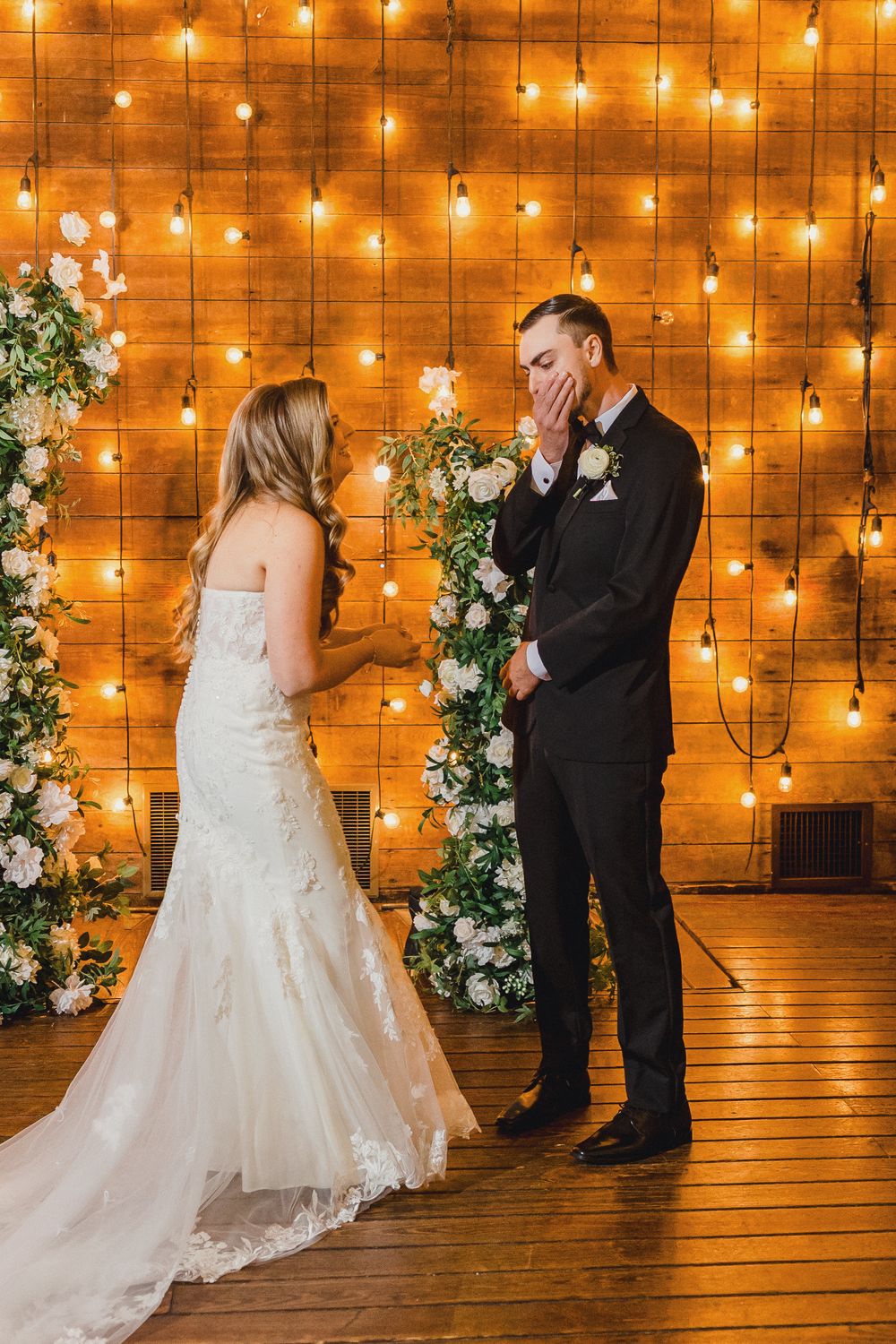 A romantic wedding scene with a couple sharing a moment against a wall of twinkling string lights and floral decorations.