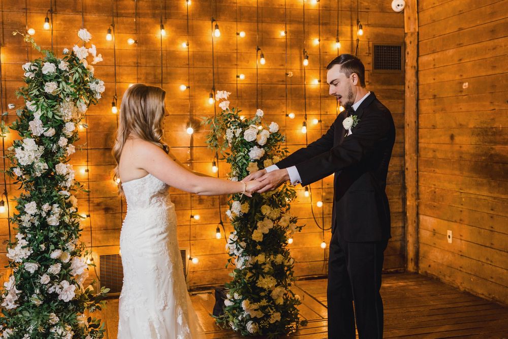 A couple exchanges vows under a wooden arch decorated with white flowers and twinkling string lights.