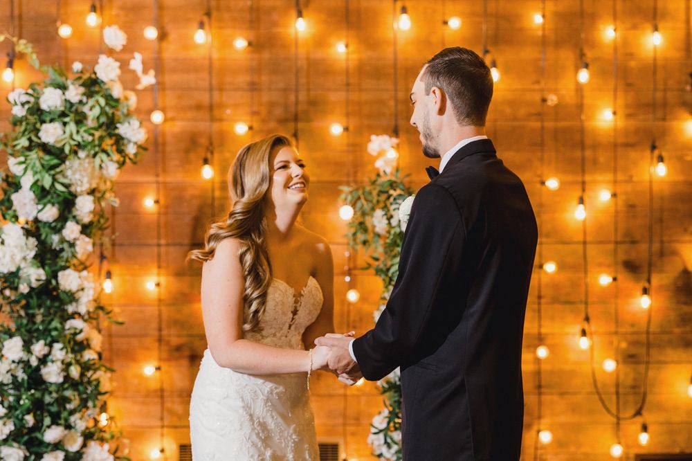 A romantic wedding ceremony setup with string lights glowing against a wooden wall backdrop and white floral arrangements.