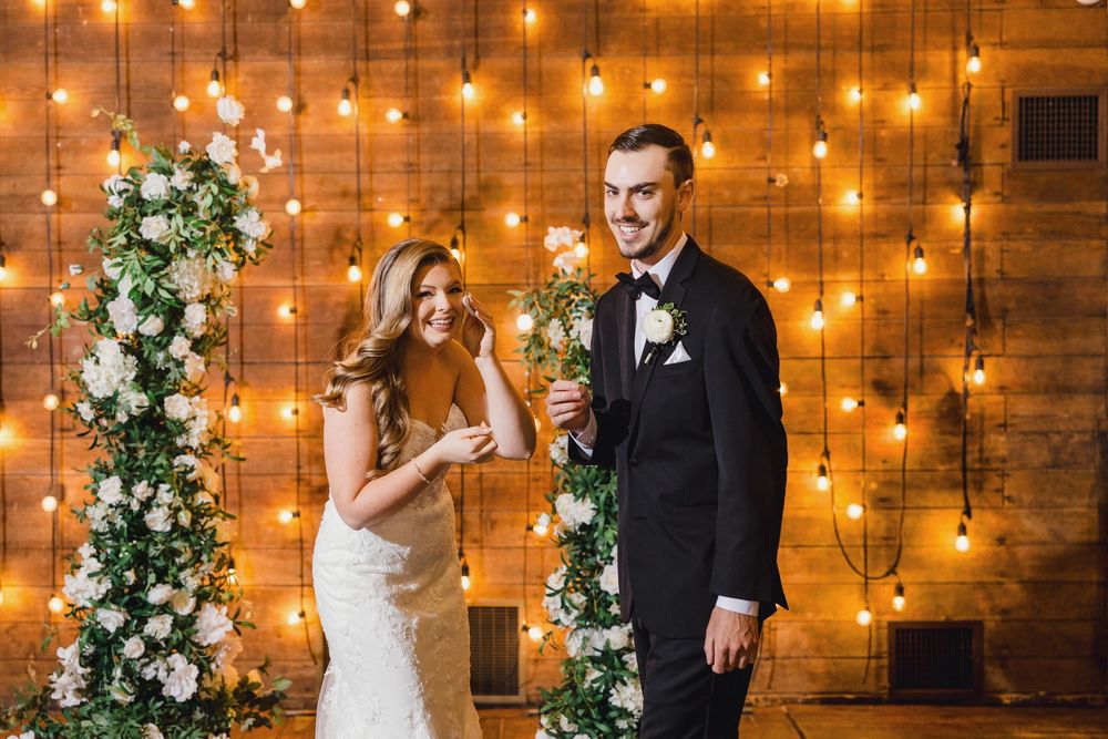 Wedding couple shares a joyful moment in front of a rustic wooden wall illuminated by string lights.