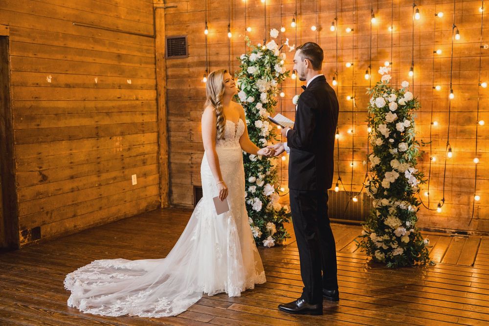 A romantic wedding ceremony setup with string lights, floral arrangements and wooden barn walls creating a warm ambiance.