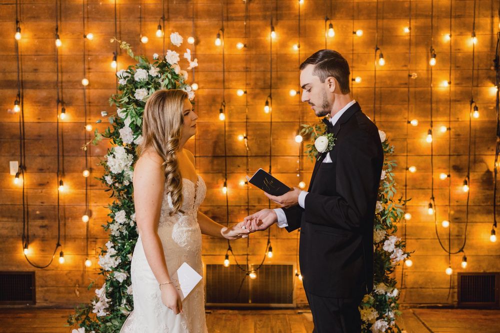 Romantic wedding ceremony moment with string lights glowing on wooden wall backdrop during evening vows.