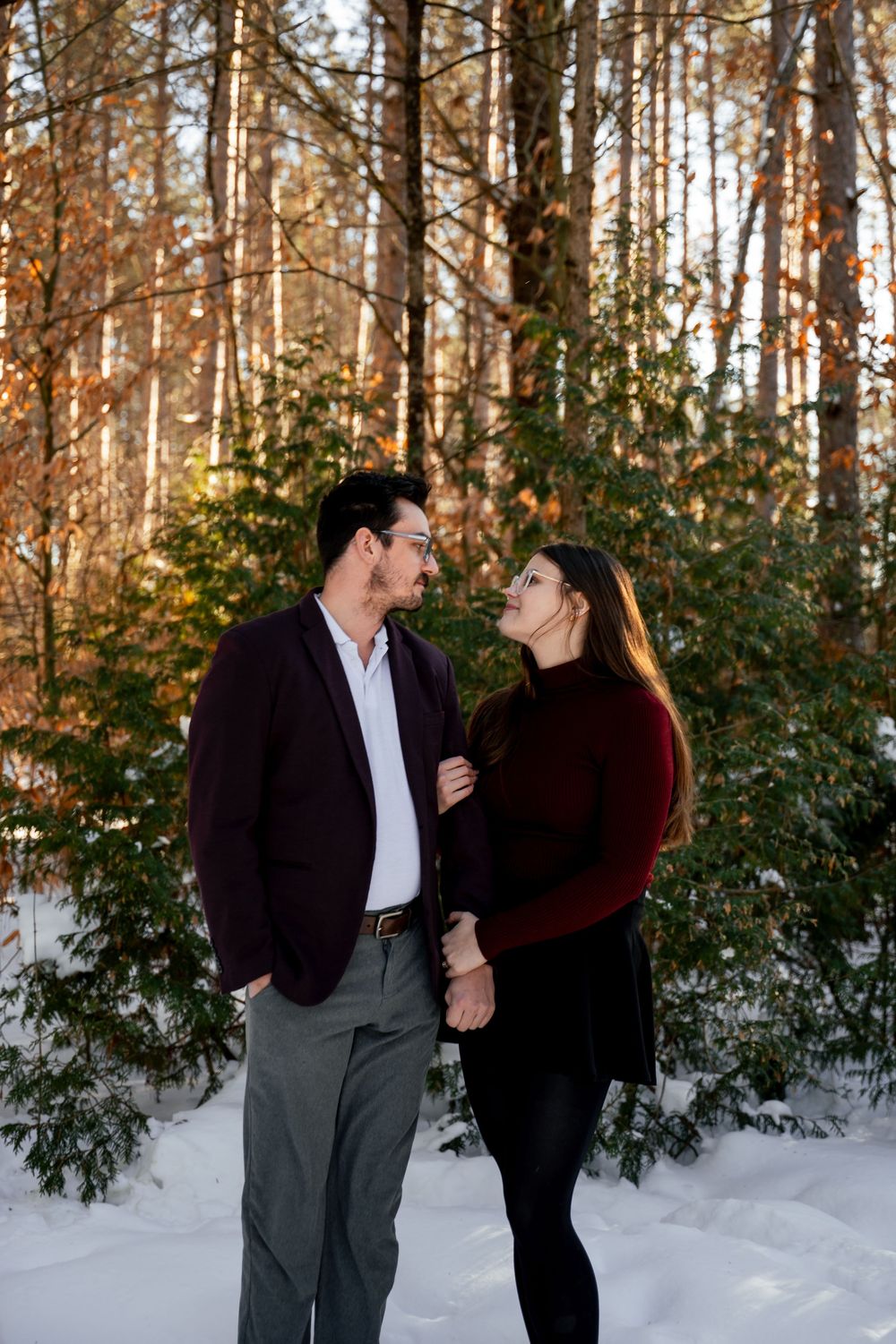 A couple shares a playful moment in a snowy forest setting with evergreen trees in the background.