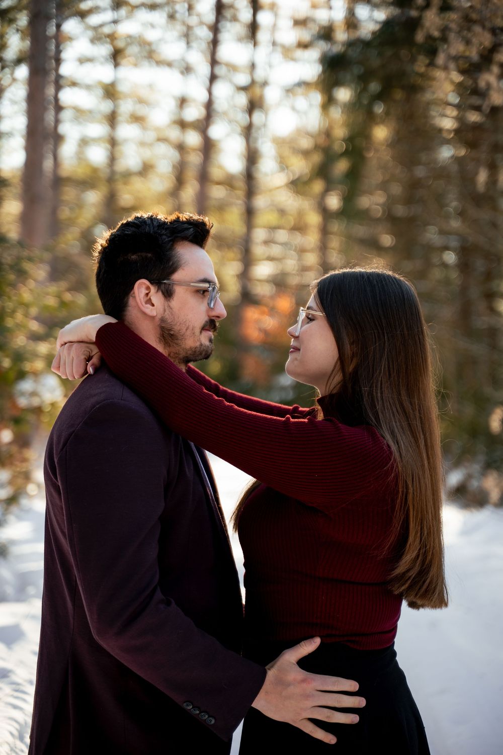 A romantic winter engagement photoshoot in a snowy forest with matching burgundy sweaters.