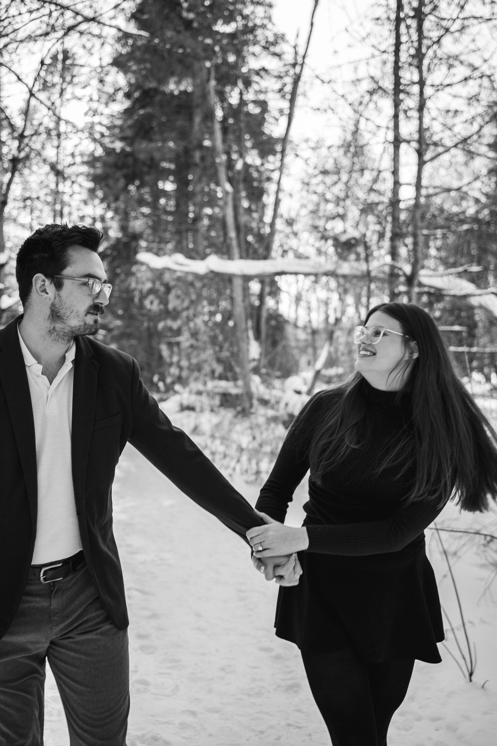 Romantic winter engagement photo of a couple walking hand in hand through a snowy forest path.