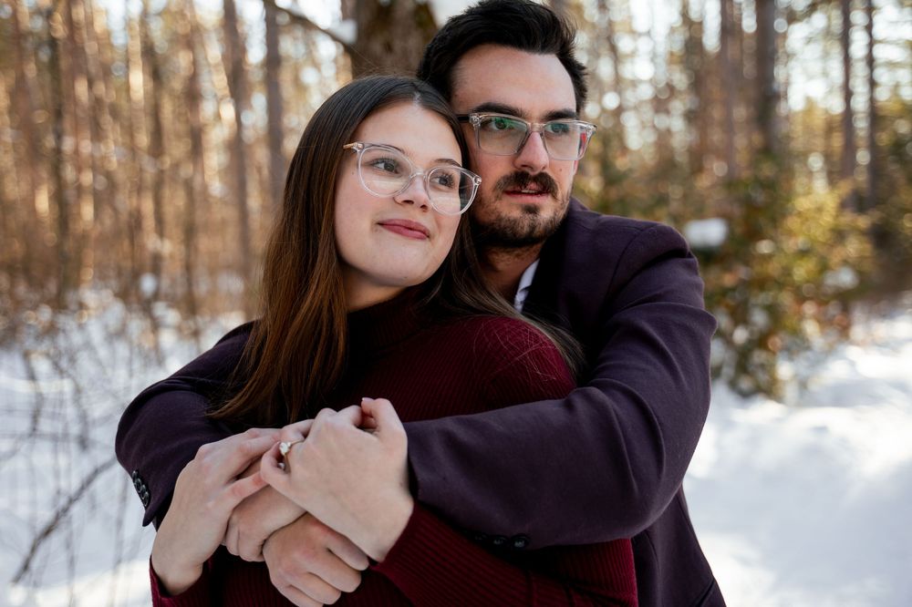 A couple embraces in a snowy winter forest setting with bare trees in the background.