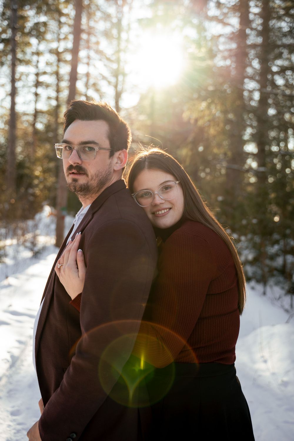 A romantic winter portrait of a couple in burgundy coats embracing in front of snowy evergreen trees at sunset.