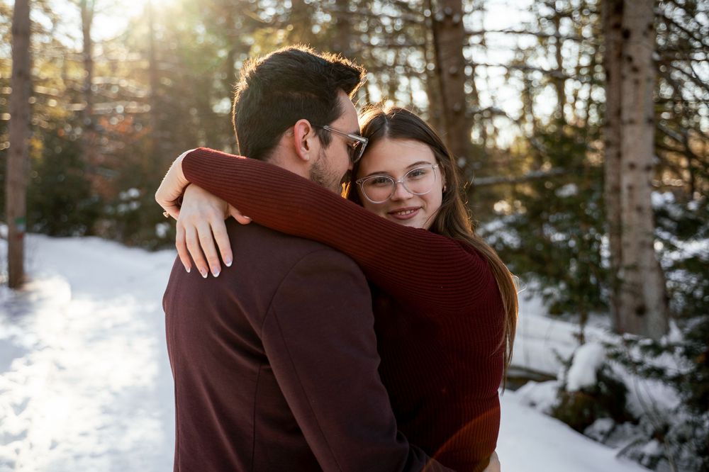 Two people embrace in a snowy winter forest setting with sunlight filtering through the trees.