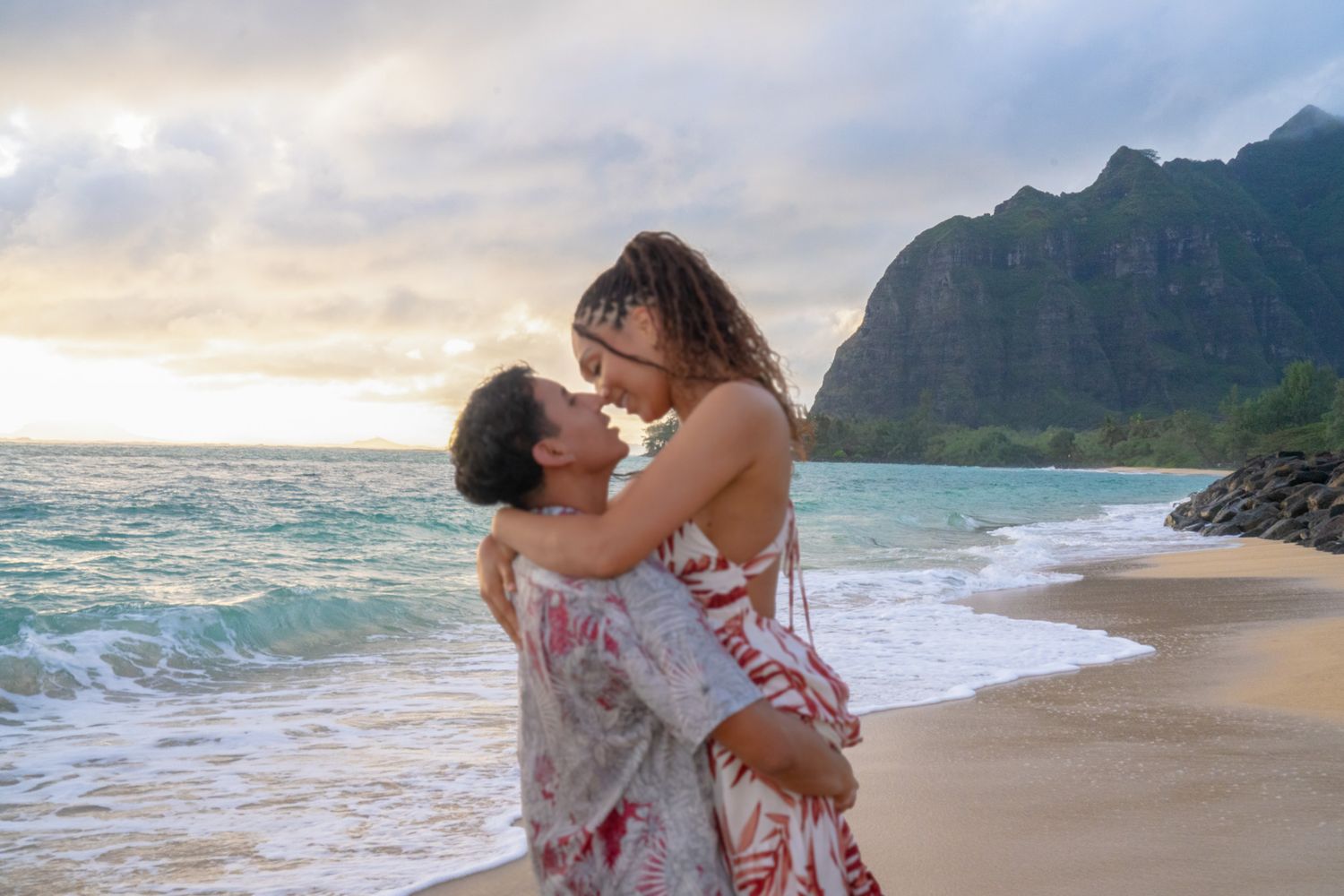 Couple in red and white outfits sharing romantic moments on a scenic beach with mountains and ocean waves at sunset.