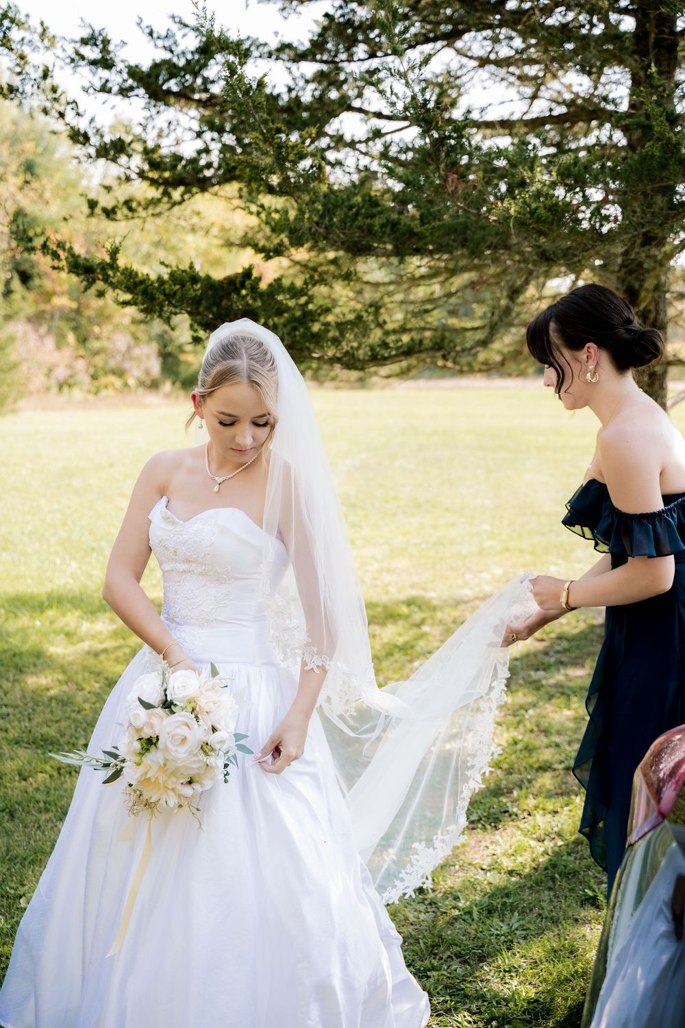 A bride in a white wedding dress is helped with her gown outdoors while holding a bouquet of flowers.