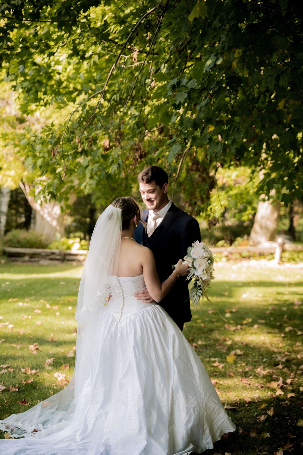 A couple in wedding attire shares intimate moments under green trees in a romantic outdoor garden setting.