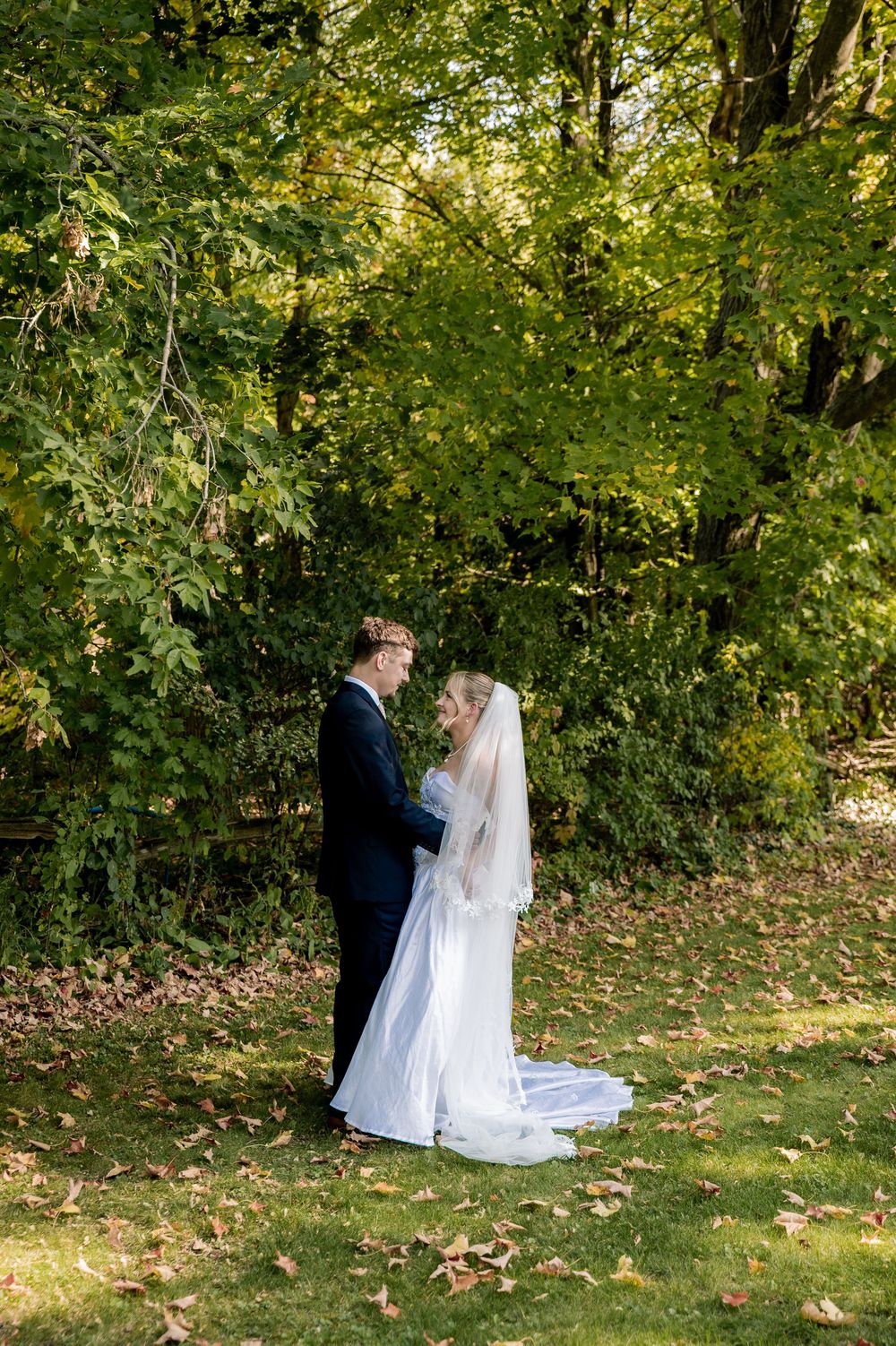 A couple shares a romantic moment in their wedding attire under lush green trees in an autumn garden setting.