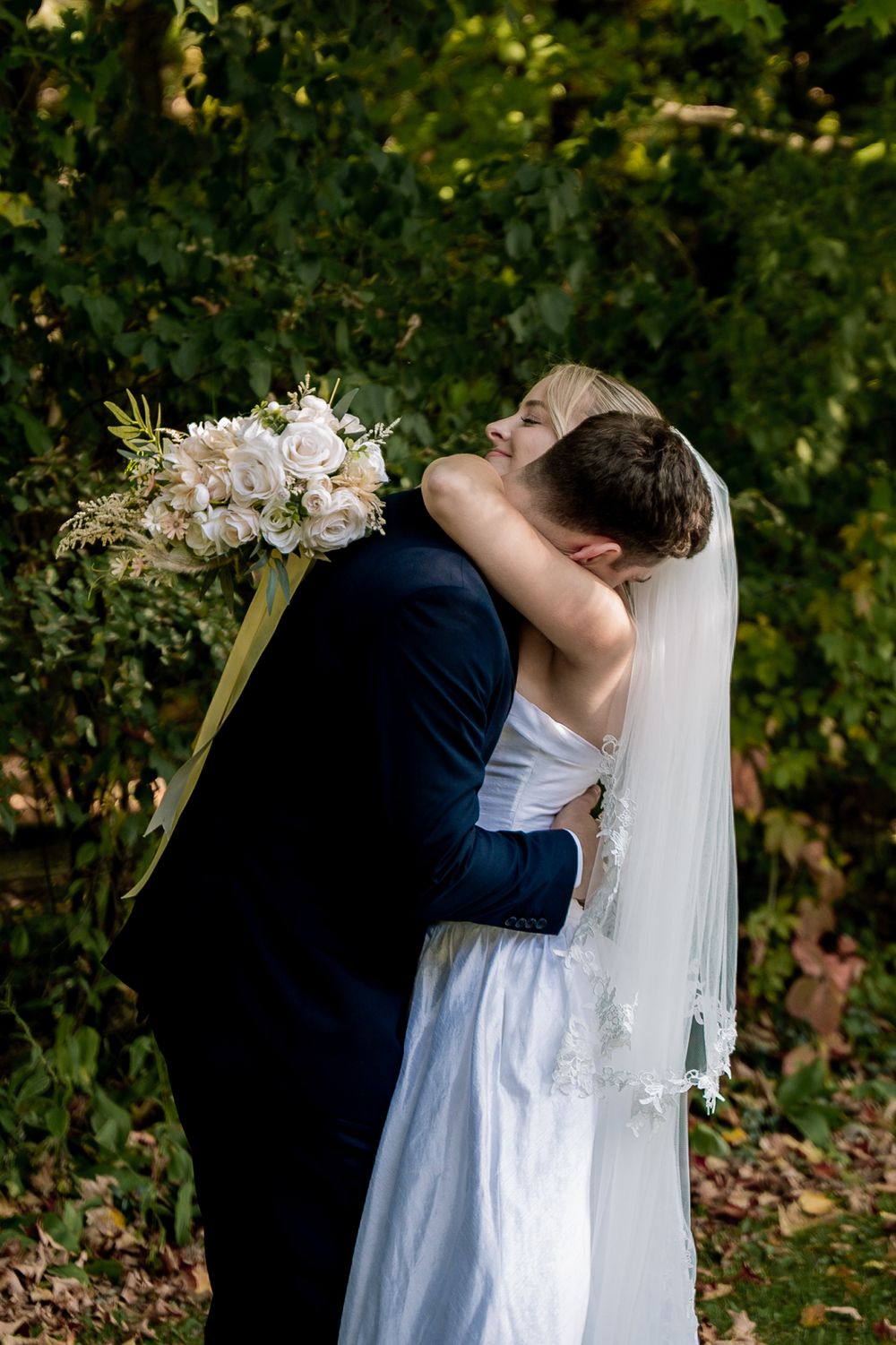A romantic wedding couple embraces in a garden setting with the bride wearing a flowing white dress and veil.