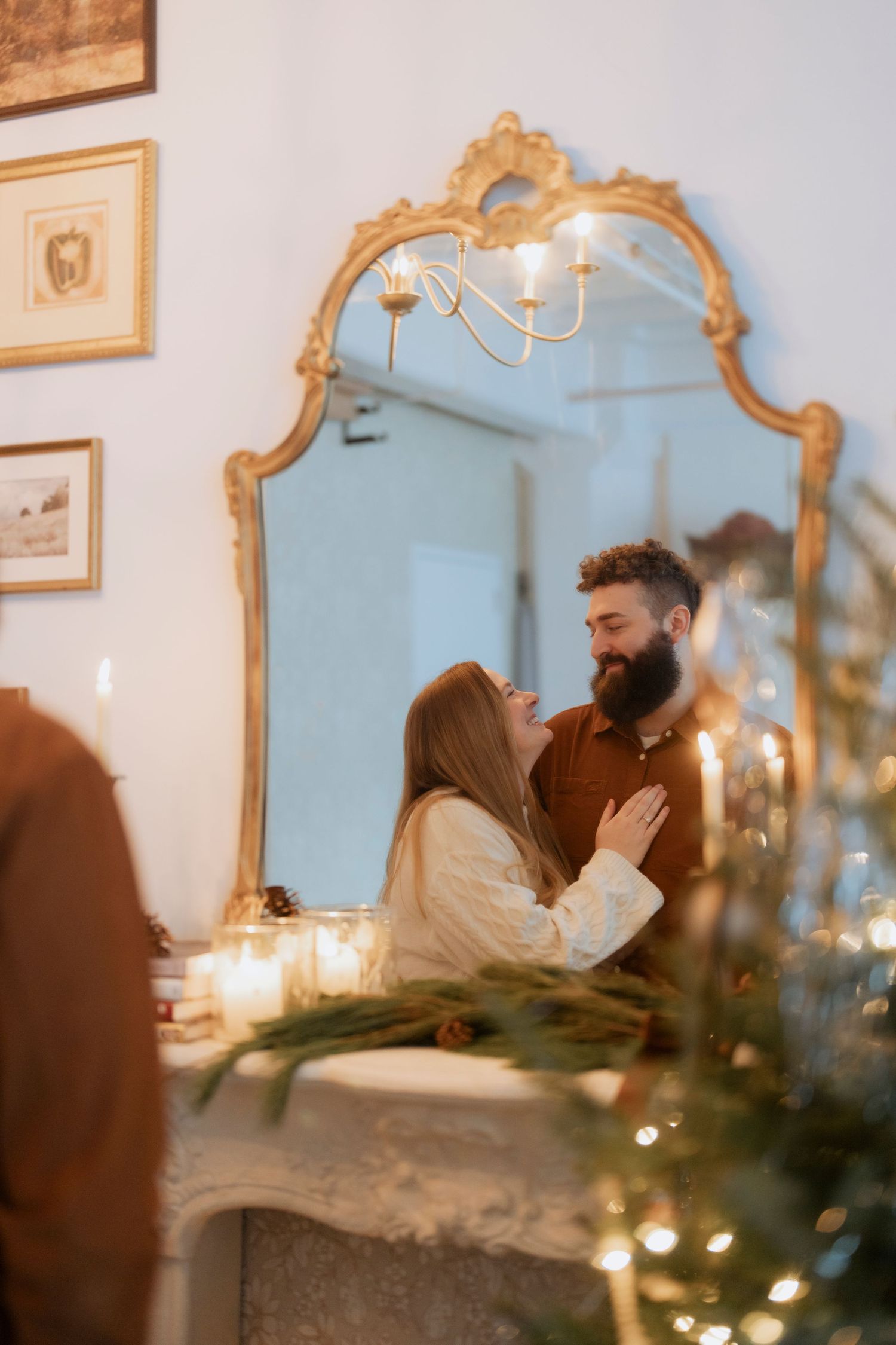 A couple shares an intimate moment reflected in an ornate gold mirror surrounded by candlelight and holiday decorations.