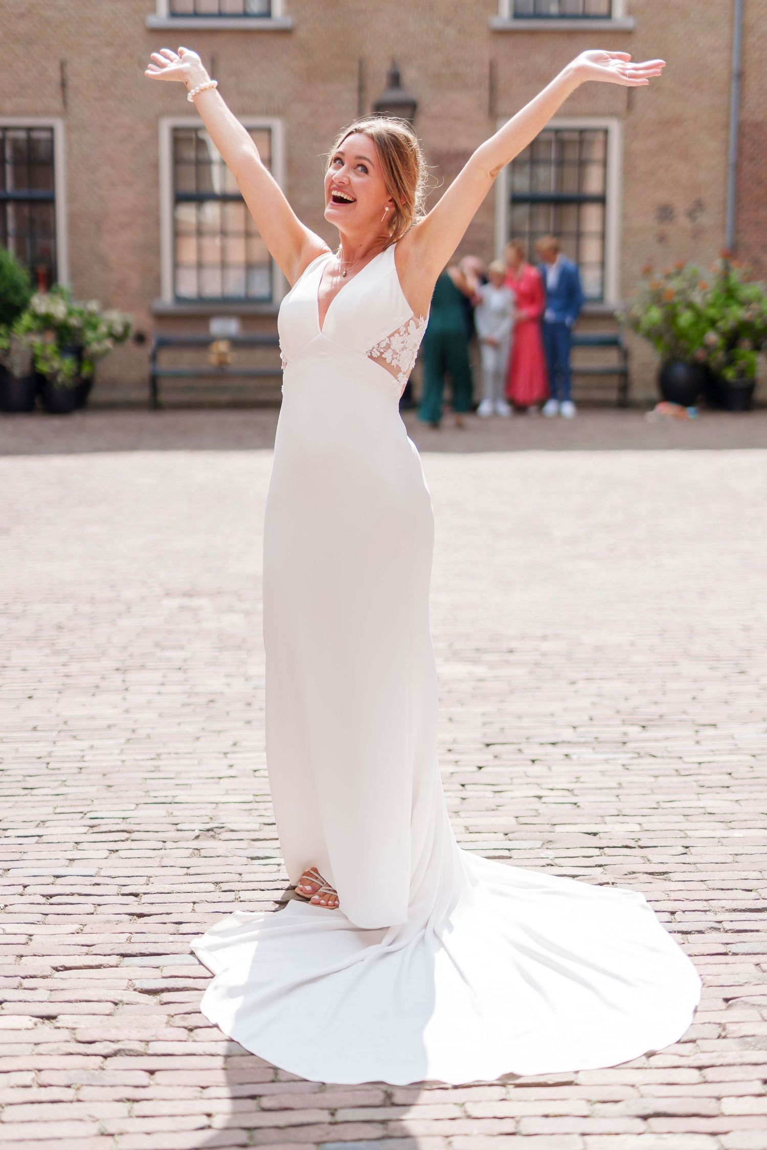 A joyful bride in a sleek white mermaid wedding dress throws her arms up in celebration on a cobblestone courtyard.