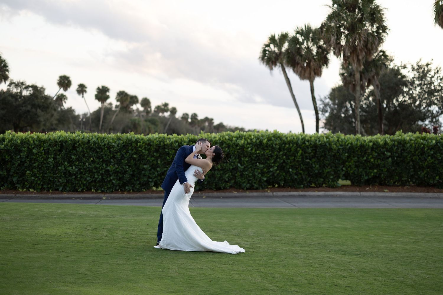 A wedding couple poses in a scenic outdoor garden setting with palm trees and lush greenery in the background.