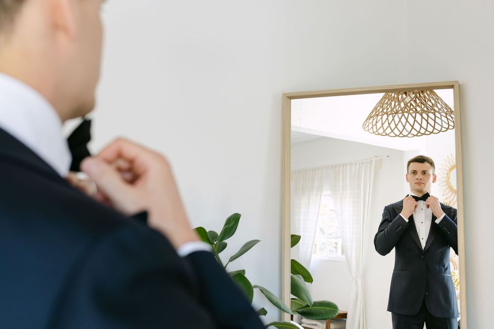 A groom adjusts a black tie while wearing a formal suit in a bright interior setting with houseplants in a venue in lisbon