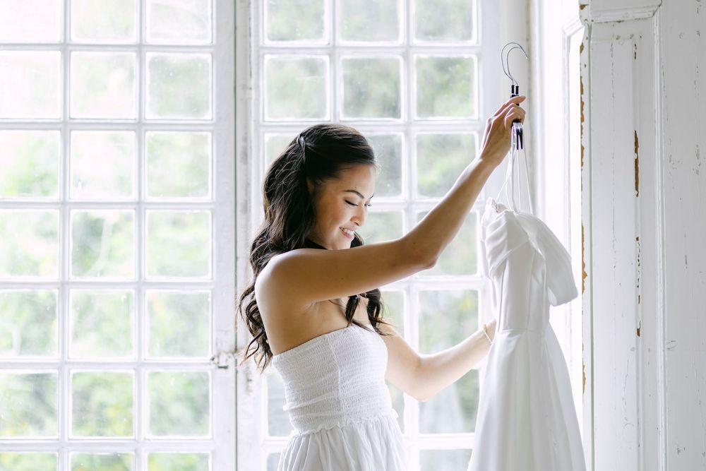 Brides admires her white wedding dress hanging by a bright window in a sunlit room during destination wedding in Portugal