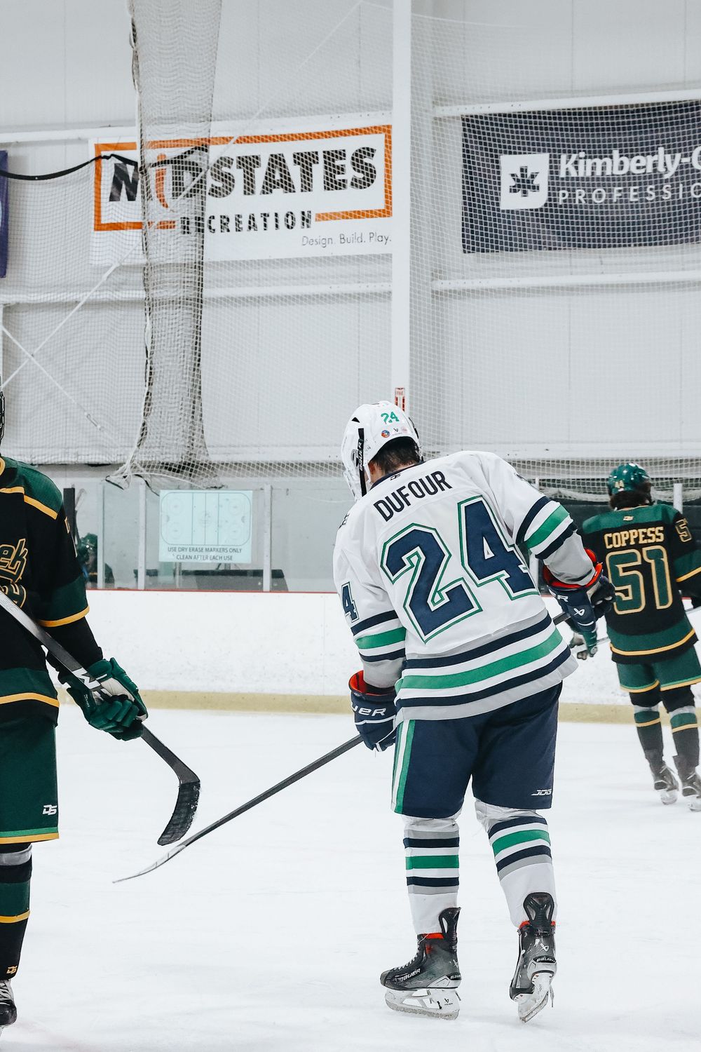 Hockey players in green and white jerseys compete during an ice hockey match on indoor rink.